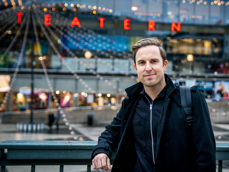 En porträttbild på Andreas. Han står och lutar sig mot ett staket. Bakom honom hänger ljuslyktor och ljusslingor och på byggnaden bakom honom står det teater med stora röda bokstäver.