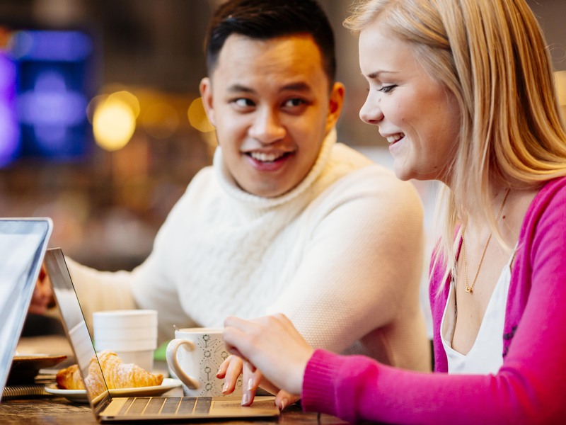 Två studenter sitter vid ett bord och samtalar. Framför dem står fika och datorer. 