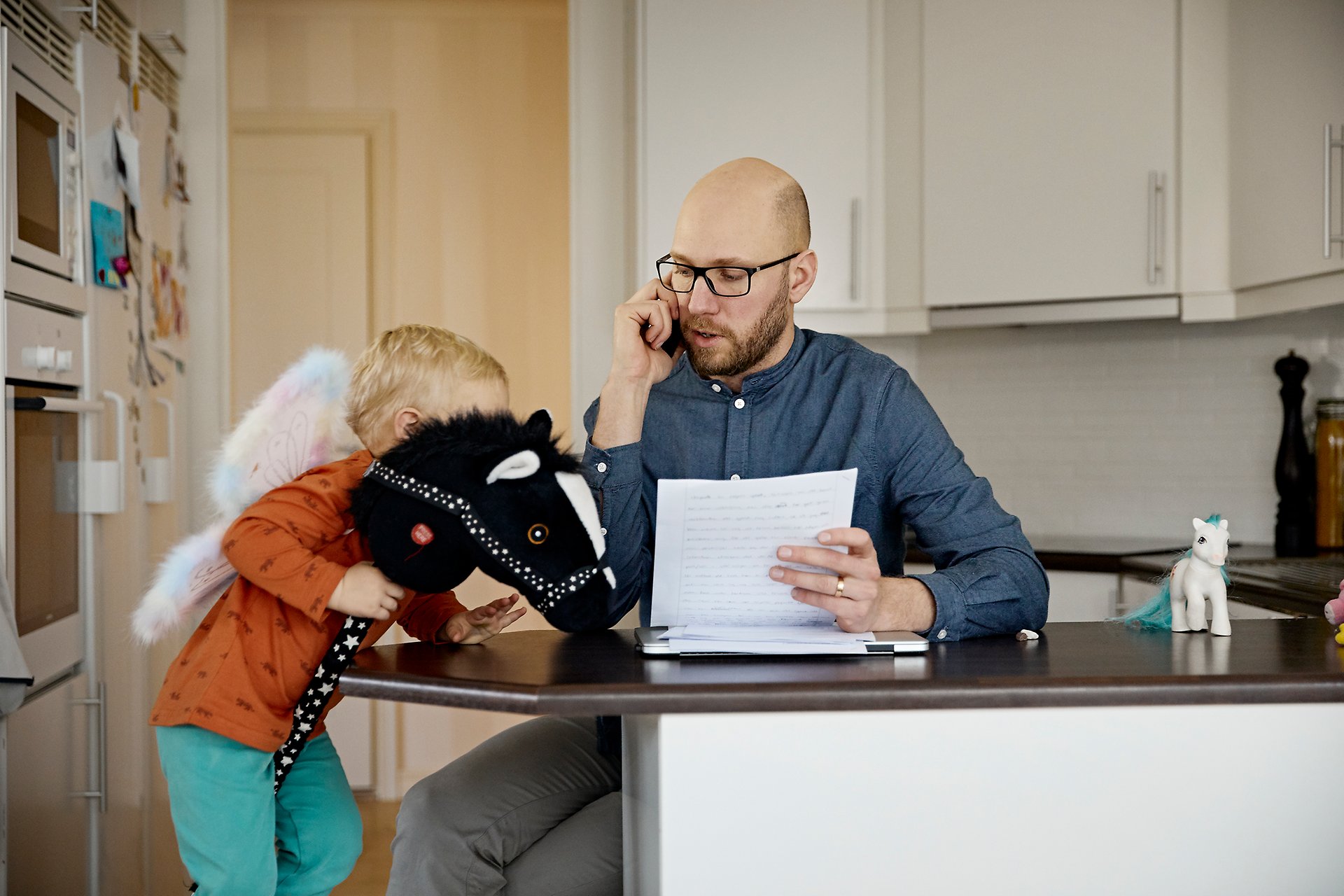 Barn med käpphäst och pappa i mobilsamtal i ett kök 