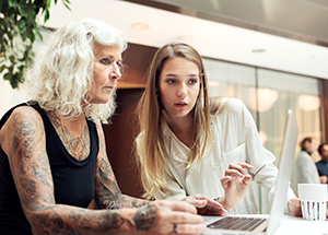 En äldre elev eller skolpersonal sitter framför en dator. En yngre kvinna står bredvid och de båda tittar på datorskärmen.