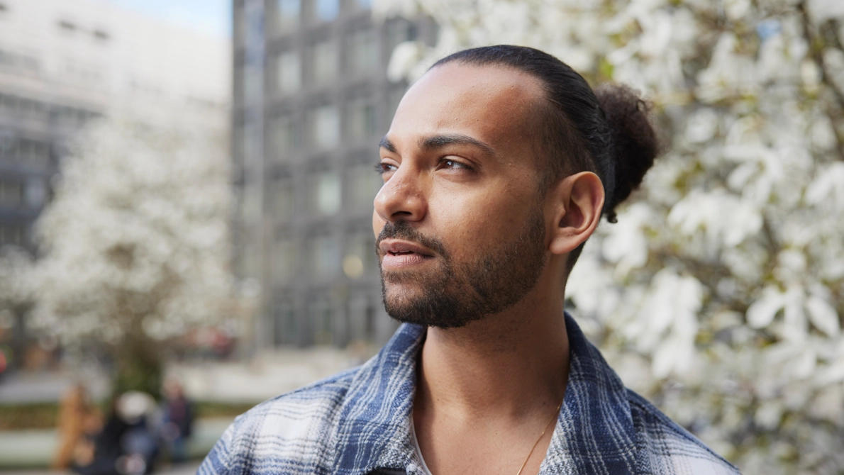 En man står och tittar bort från kameran. I bakgrunden syns ett höghus och blommande träd.