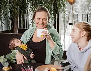 En vuxen kvinna håller i ett litet barn och samtidigt en kopp som hon ska dricka ur. Bredvid henne sitter ett äldre barn, en flicka. 