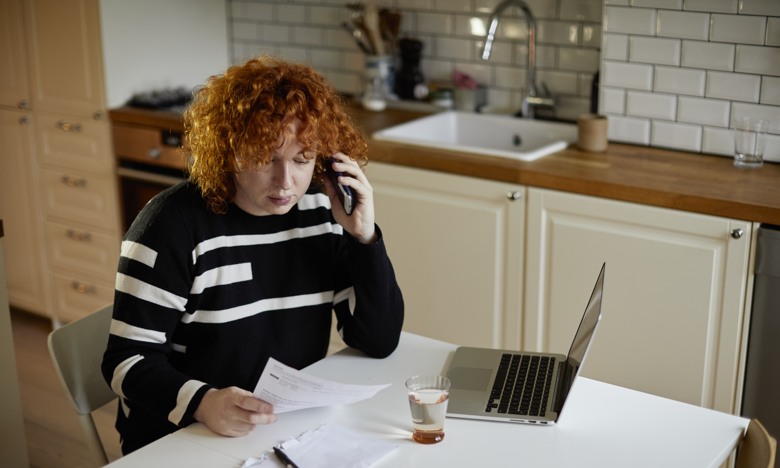 Rödhårig kvinna sittandes vid ett bord med telefonen vid örat och ett brev och en dator framför sig