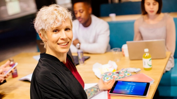 Gråhårig kvinna tittar in i kameran och ler. Bakom henne sitter en man och kvinna och arbetar på sina datorer.